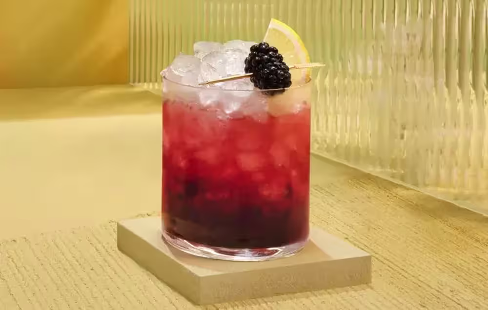 Ein mit Crushed Ice und einem rot-violetten Getränk gefülltes Glas, garniert mit einer Brombeere und einem Zitronenschnitz auf einem Zahnstocher, steht auf einem quadratischen, hellen Untersetzer vor einem gelben, strukturierten Hintergrund.