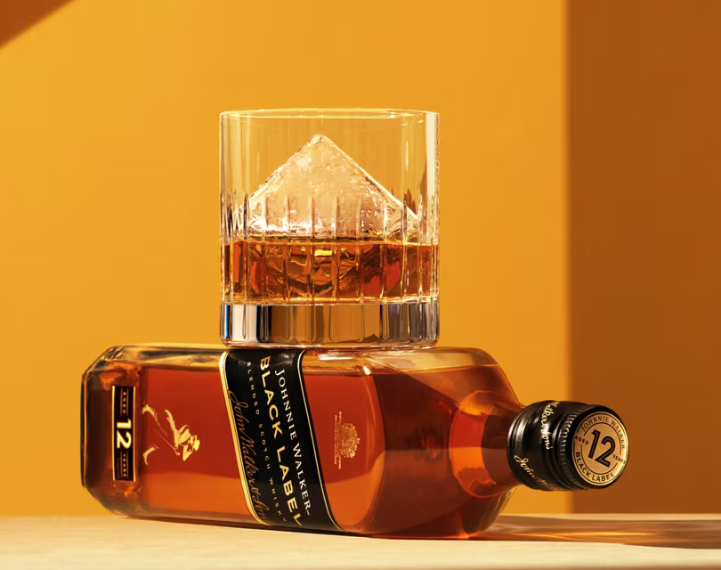A glass of whisky with a large diamond-shaped ice cube sits on top of a tilted bottle of Johnnie Walker Black Label 12-year-old whisky against a yellow background.
