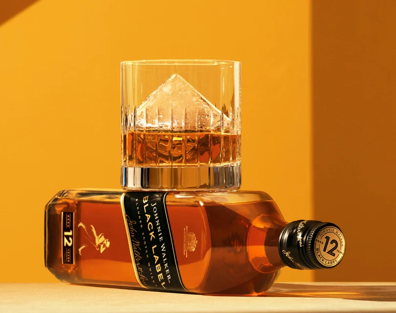 A glass of whisky with a large ice cube sits on top of a tilted bottle of Johnnie Walker Black Label against a warm orange background. The bottle is positioned horizontally, showcasing the label.