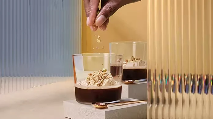 A hand sprinkles chopped nuts onto a glass of dark dessert topped with cream. Another identical dessert sits beside it on a white block with two small golden spoons nearby. The background features textured panels.