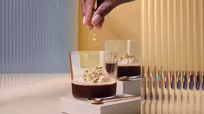 Une main saupoudre des noix hachées sur un verre de dessert noir garni de crème. Un autre dessert identique est posé à côté, sur un bloc blanc, avec deux petites cuillères dorées à proximité. L'arrière-plan présente des panneaux texturés.