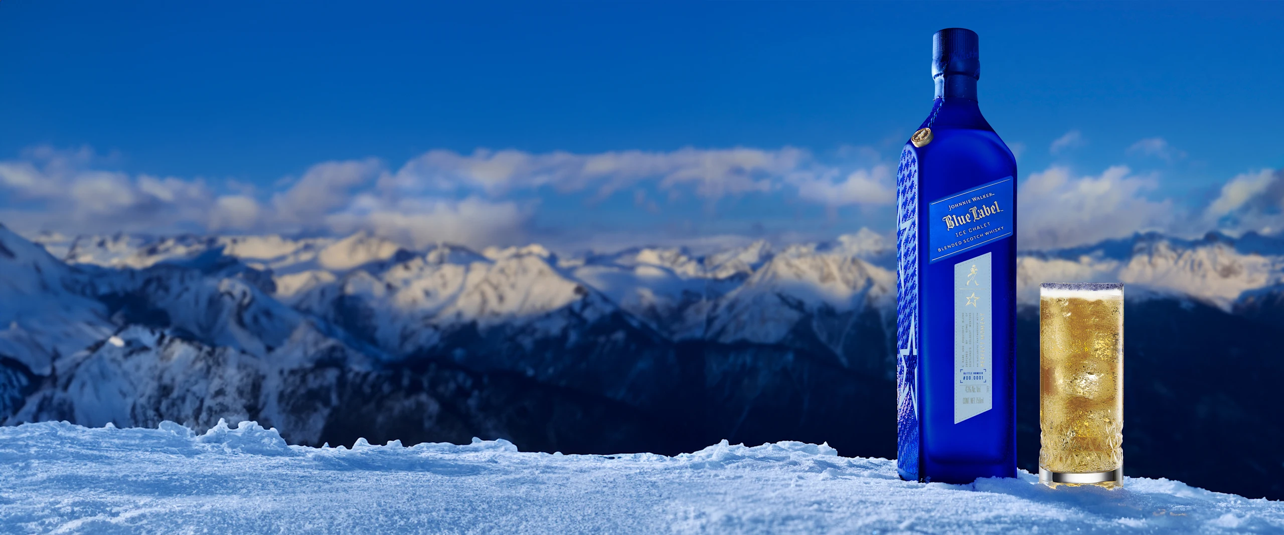 A blue bottle of Johnnie Walker Blue Label Ice Chalet and a glass filled with a golden beverage placed on a snowy surface with a backdrop of snow-covered mountains under a clear blue sky.