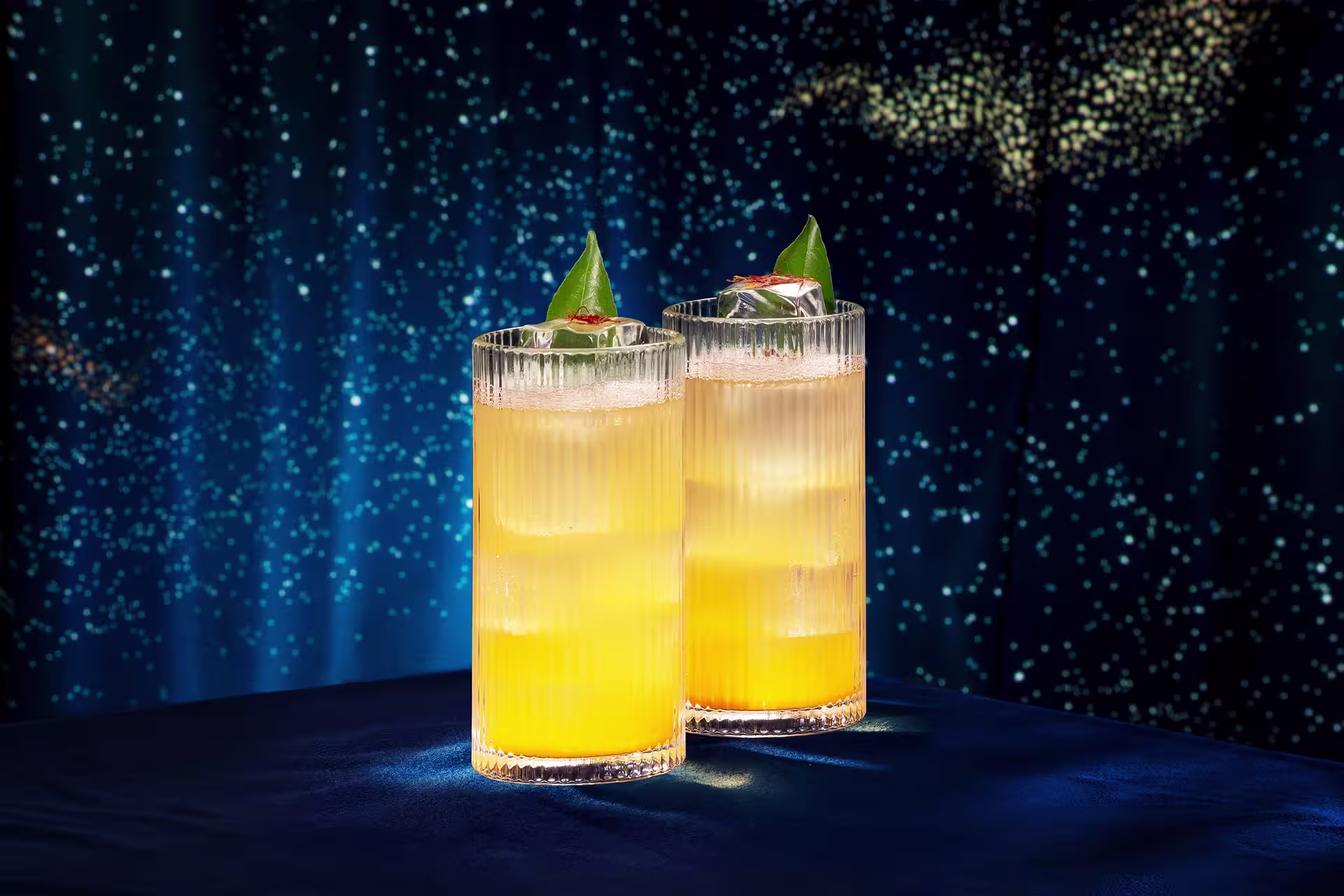 Two tall, ribbed glasses filled with a yellow-orange cocktail, garnished with green leaves and ice, are placed on a dark blue surface with a starry, night sky background.