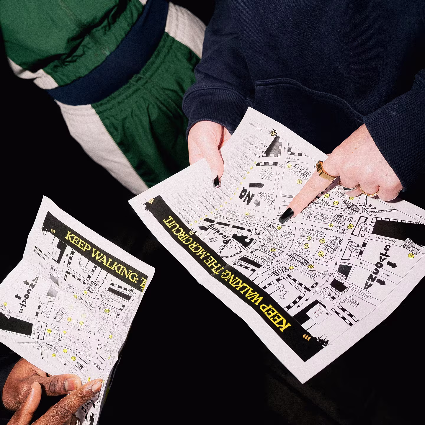 Two people holding and examining printed maps labelled KEEP WALKING, THE PATH CUT.