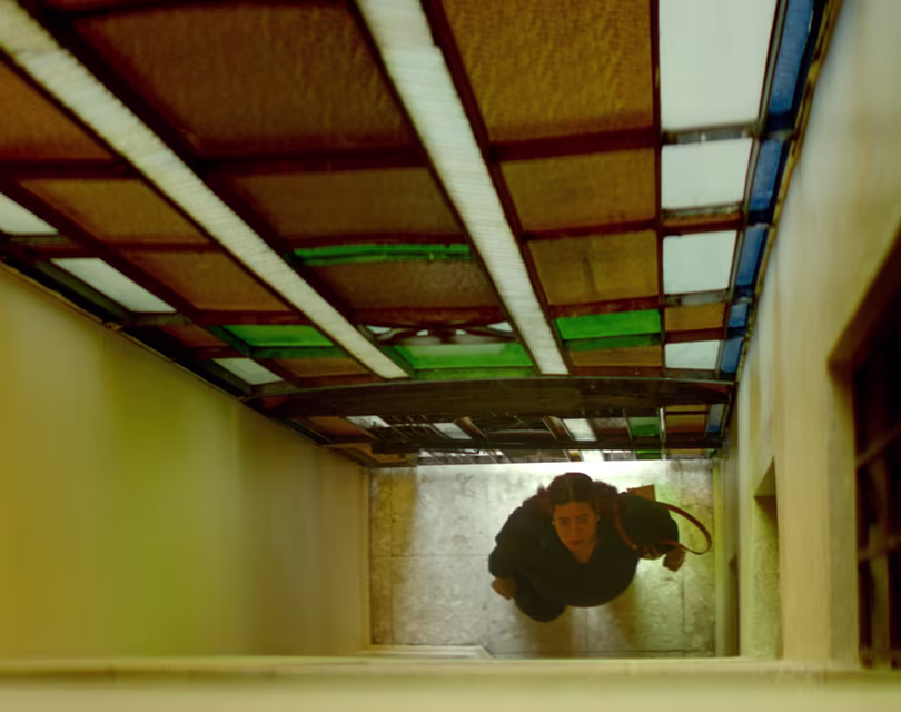 Desde arriba se ve a una persona subiendo unas escaleras estrechas con una bolsa en la mano. El pasillo tiene un techo de vidrio con tonos marrones y verdes que proyectan una luz colorida. Las paredes son amarillentas, lo que crea un ambiente cálido.