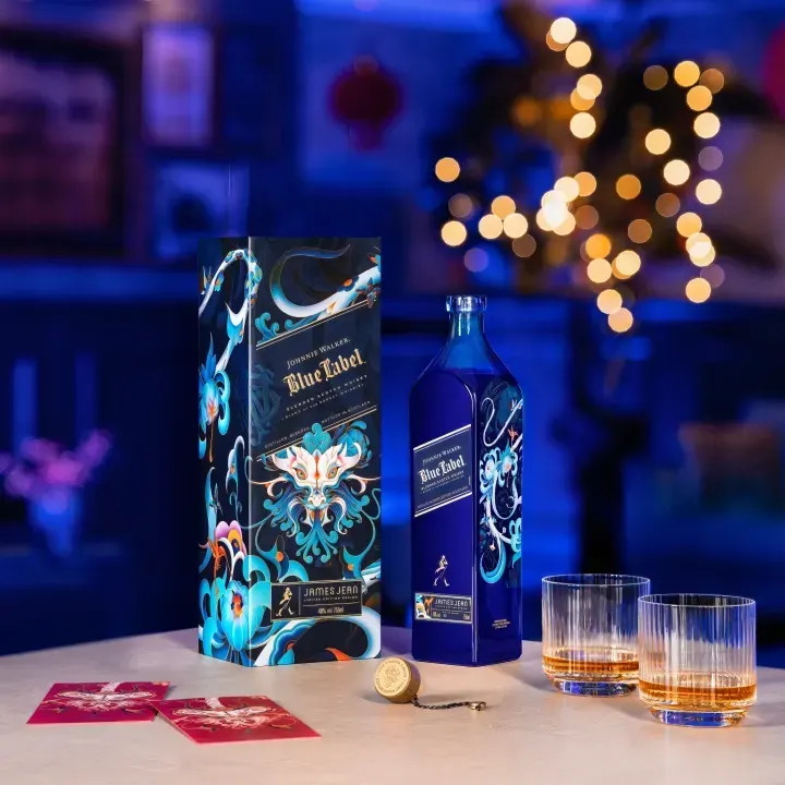 A bottle of Blue Label Limited Edition Design Year of the Dragon whiskey and its ornate box sit on a table alongside two filled glasses and a gold seal. In the background, there are blurred lights, suggesting a festive ambiance.