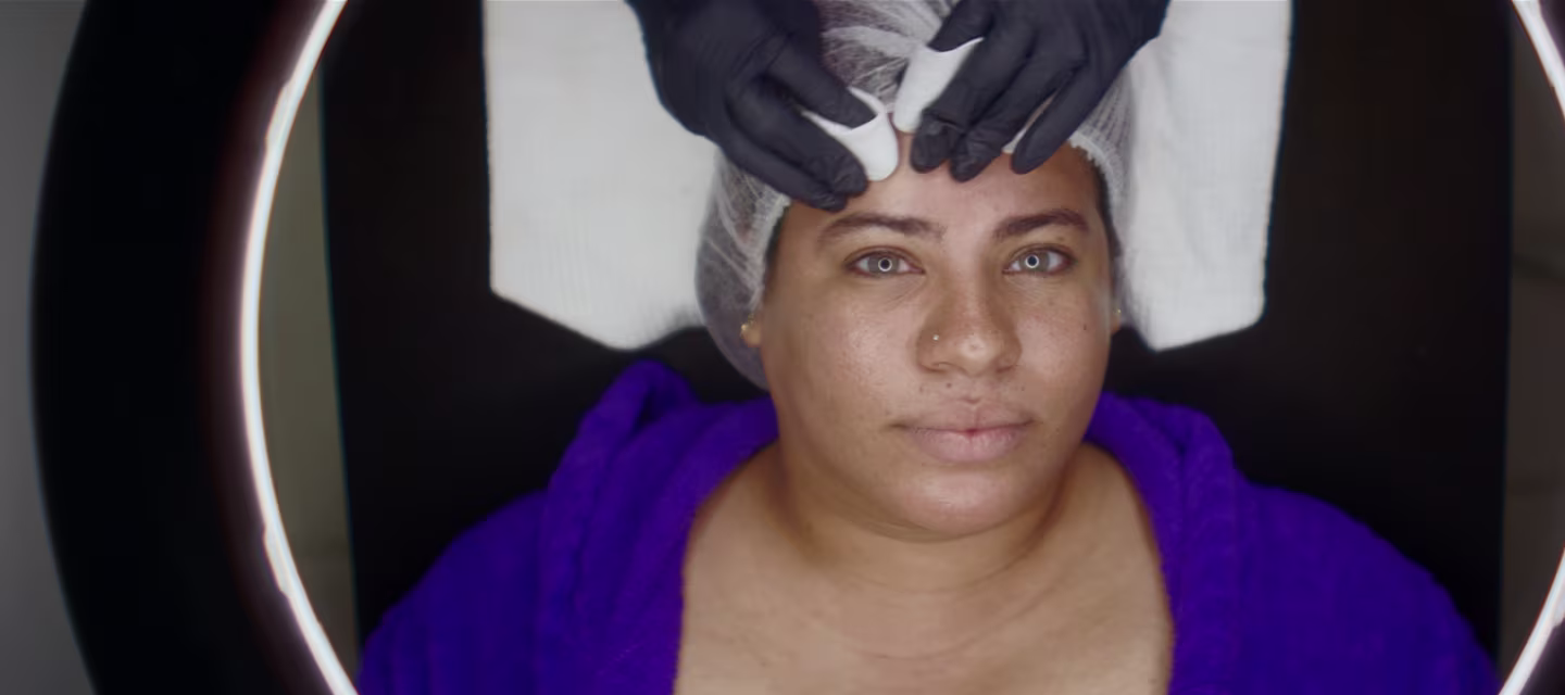 Una persona con expresión serena yace bajo una luz circular mientras recibe un tratamiento facial. Lleva una túnica violeta y una redecilla para el pelo, y con las manos enguantadas se aplica suavemente un producto o un masaje en la frente.