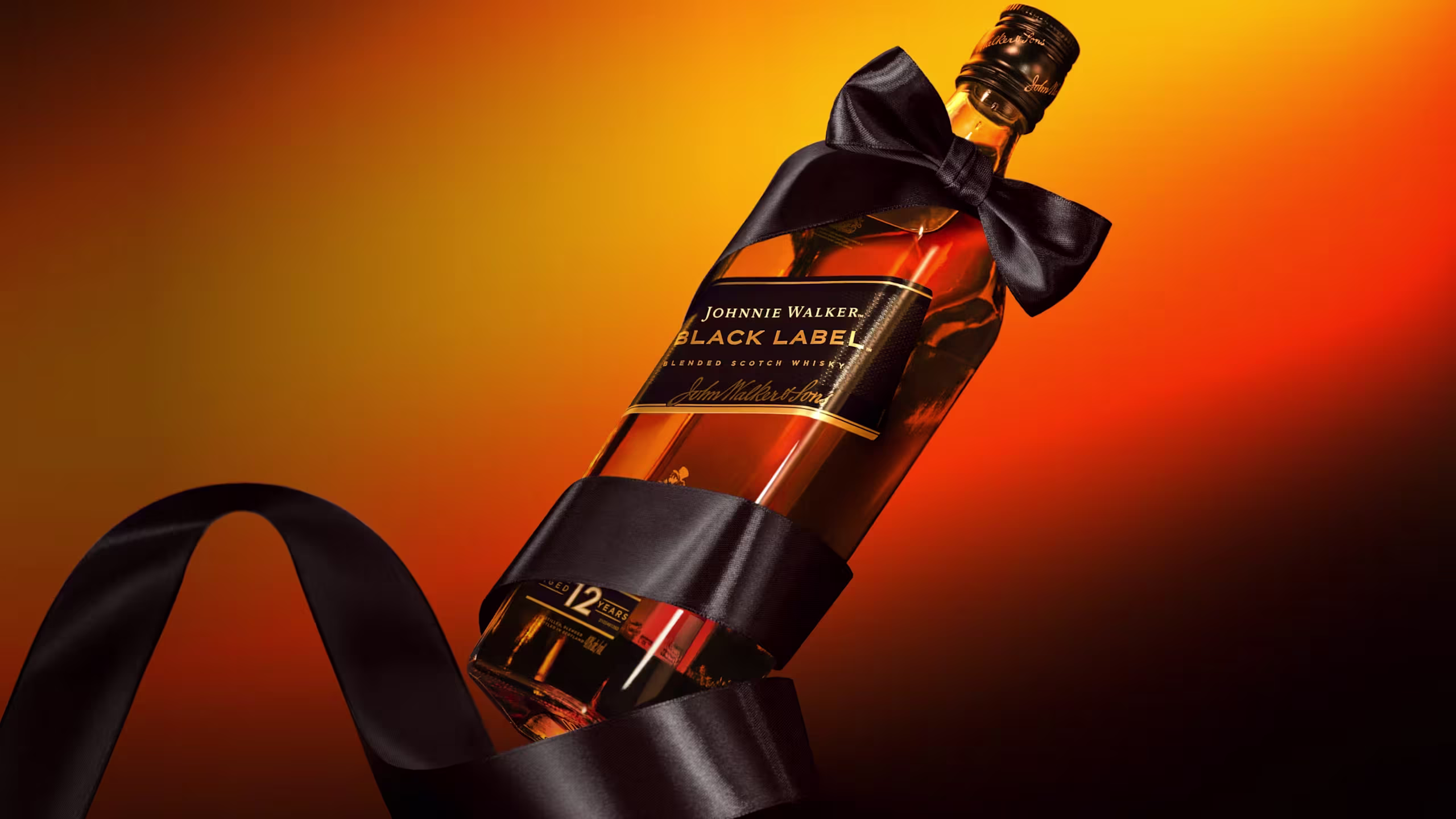 Láhev whisky Johnnie Walker Black Label omotaná černou saténovou stuhou s mašlí na zářivě oranžovém a žlutém gradientním pozadí.