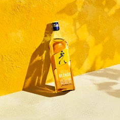  一瓶尊尼獲加金色威士忌矗立在充滿活力的黃色背景下。該標籤具有標誌性的步行者標誌。正午的陽光投下陰影，增加了場景的深度和對比。