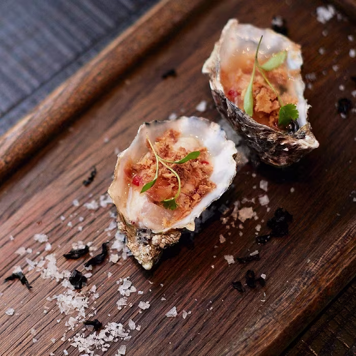 Two garnished oysters served on a wooden board with sea salt and herbs sprinkled around them.