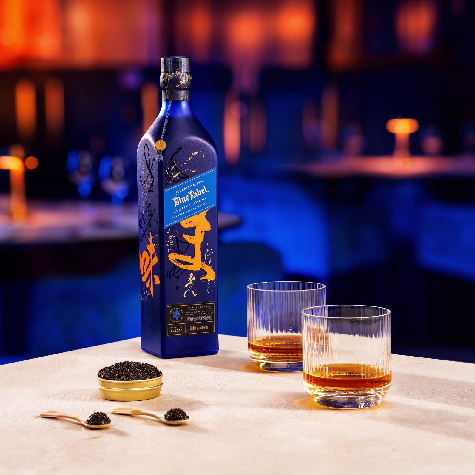 Một chai rượu whisky màu xanh đặt trên bàn cạnh hai chiếc ly đựng đầy thứ đồ uống màu hổ phách. Một hộp trứng cá muối nhỏ được mở gần đó. Bối cảnh là một quầy bar được chiếu sáng nhẹ nhàng.