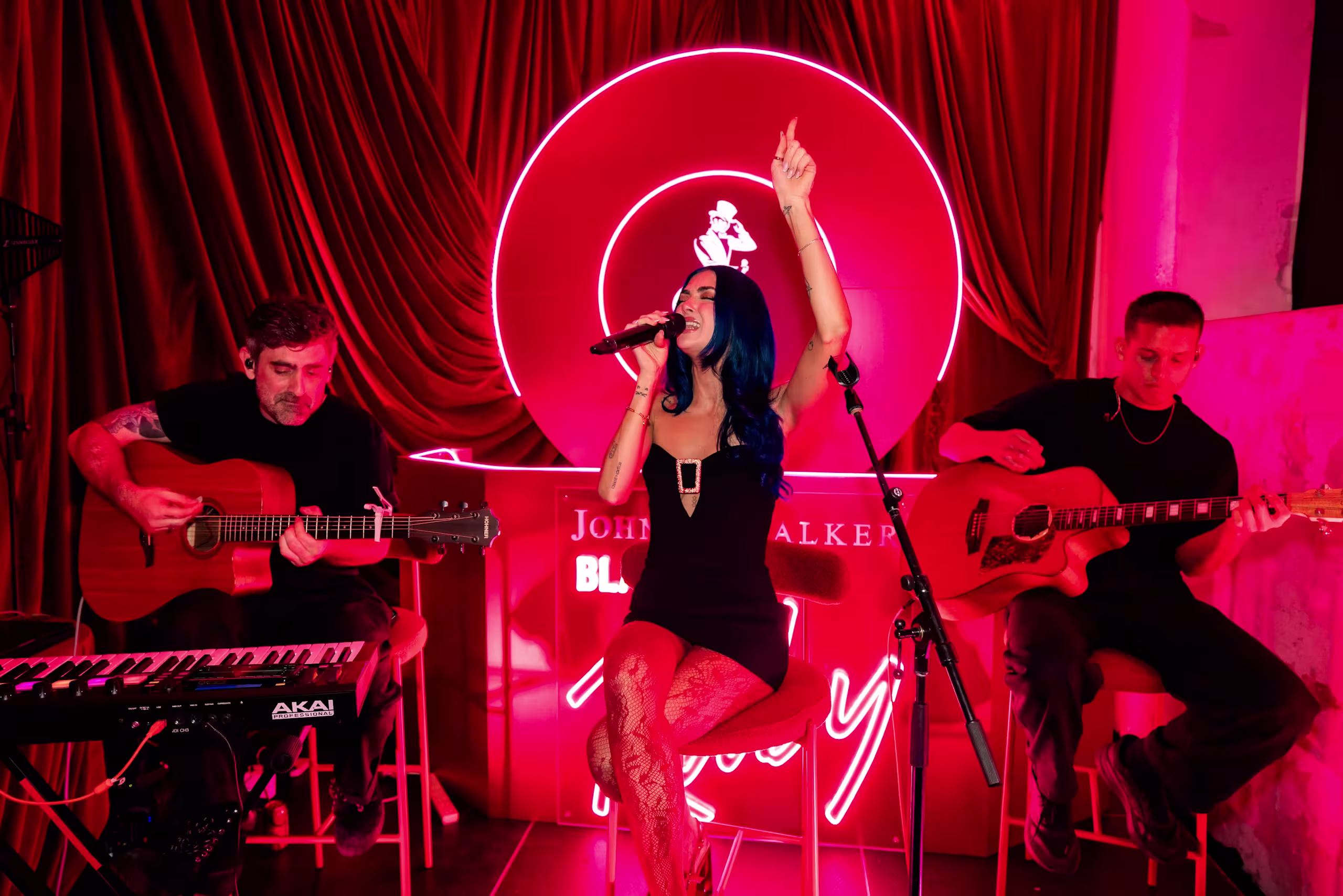 A woman sings passionately into a microphone on stage, raising one arm, while two men play acoustic guitars beside her. The scene is bathed in red light with a neon sign glowing in the background.
