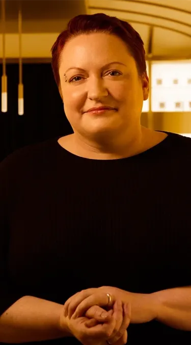 Dr. Emma Walker with short hair standing in a warmly lit room full of shelves lined with bottles. They are wearing a black top, with hands clasped. Tables with glassware are visible in the background.