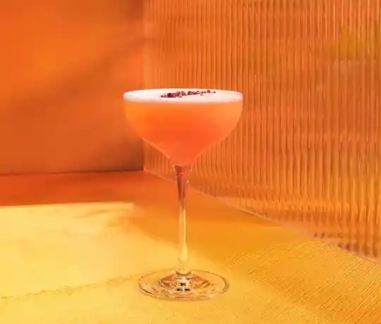 Un coctel rosado en una copa con tallo descansa sobre un mantel naranja.