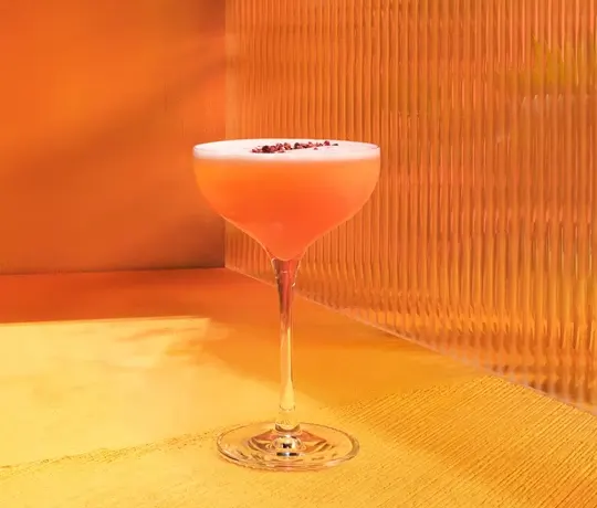 Un verre coupé rempli d'un cocktail rose-orange vif, surmonté d'une couche de mousse et d'une petite garniture rouge, repose sur une surface jaune contre un mur orange texturé.