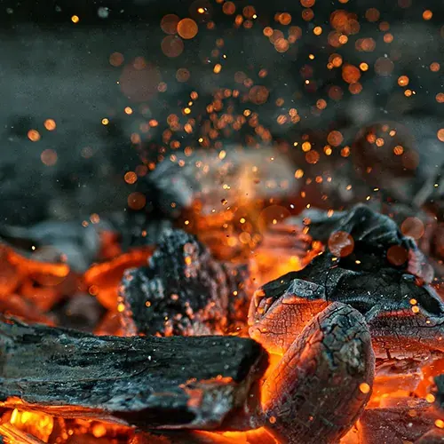 Nahaufnahme von glühender Glut und verkohlten Holzstücken in einem lebhaften Feuer, wobei leuchtend orange und rote Funken vor einem dunklen Hintergrund nach oben fliegen und eine warme und dynamische Szene erzeugen.