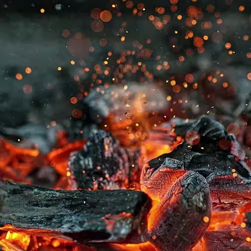  Close-up of glowing embers and charred pieces of wood in a vibrant fire, with bright orange and red sparks flying upwards against a dark background, creating a warm and dynamic scene.