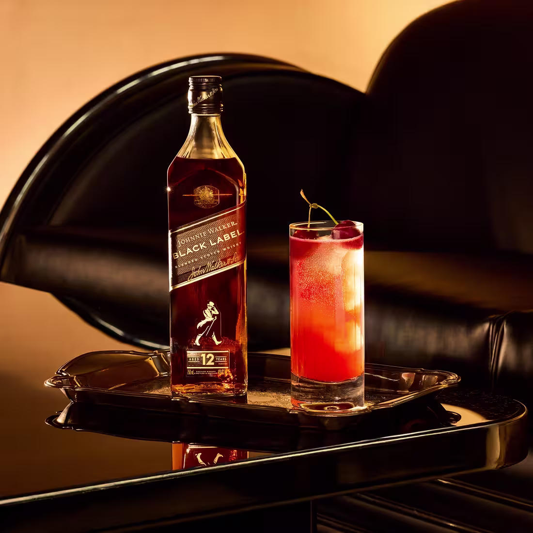 Una botella de whisky Johnnie Walker Black Label y un cóctel rojo alto adornado con un toque de cítrico se encuentran en una bandeja reflectante sobre una mesa negra brillante, con un sofá de cuero negro en el fondo.