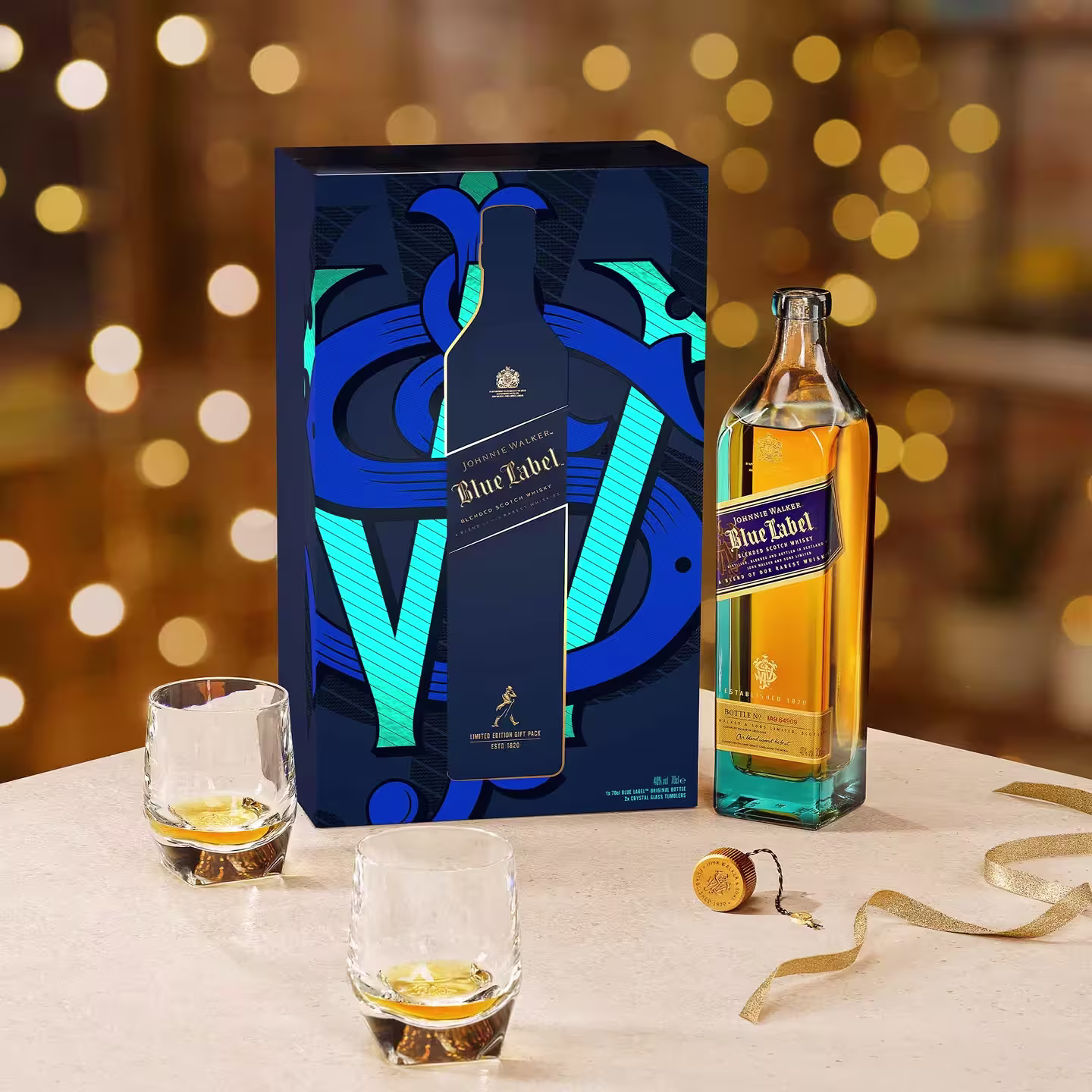 A bottle of Johnnie Walker Blue Label whisky with its decorative box, two glasses of whisky with ice, and a cork shown on a light-toned table. Warm, golden bokeh lights appear in the blurred background.