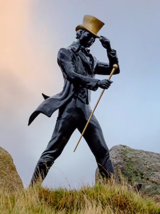 Statue of a man in motion, wearing a top hat and holding a cane, set outdoors on grass with large rocks and a cloudy sky in the background.