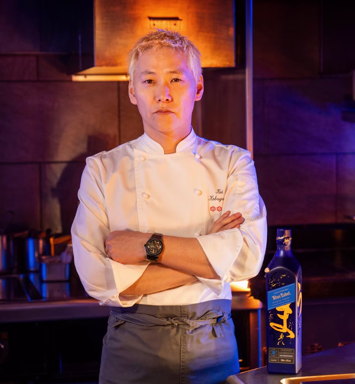 Chef KEI KOBAYASHI de jaqueta branca está com os braços cruzados em uma cozinha mal iluminada. Um Johnnie Walker Blue Label Elusive Umami com detalhes dourados está no balcão ao lado dele.