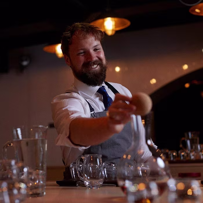 A smiling bartender in an apron serves a drink behind a bar, surrounded by glassware under warm lights.