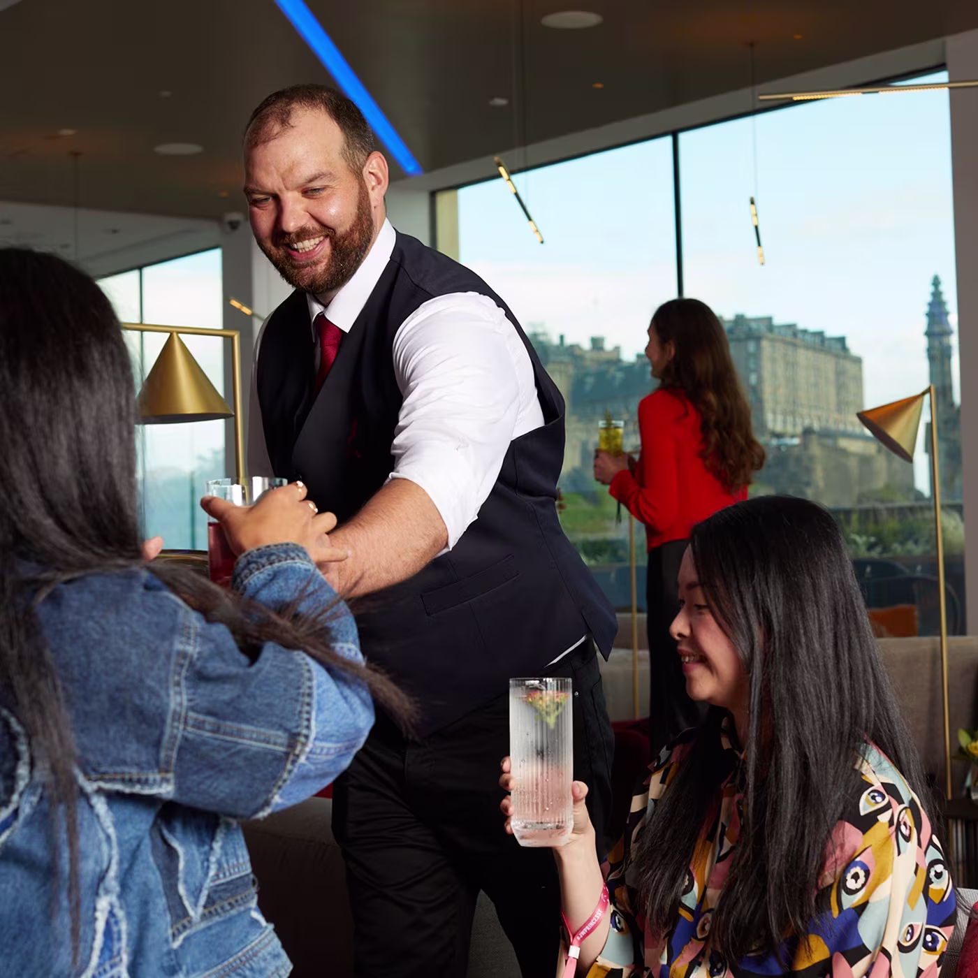 A smiling man in a vest serves drinks to two women in a modern lounge with large windows and city views.