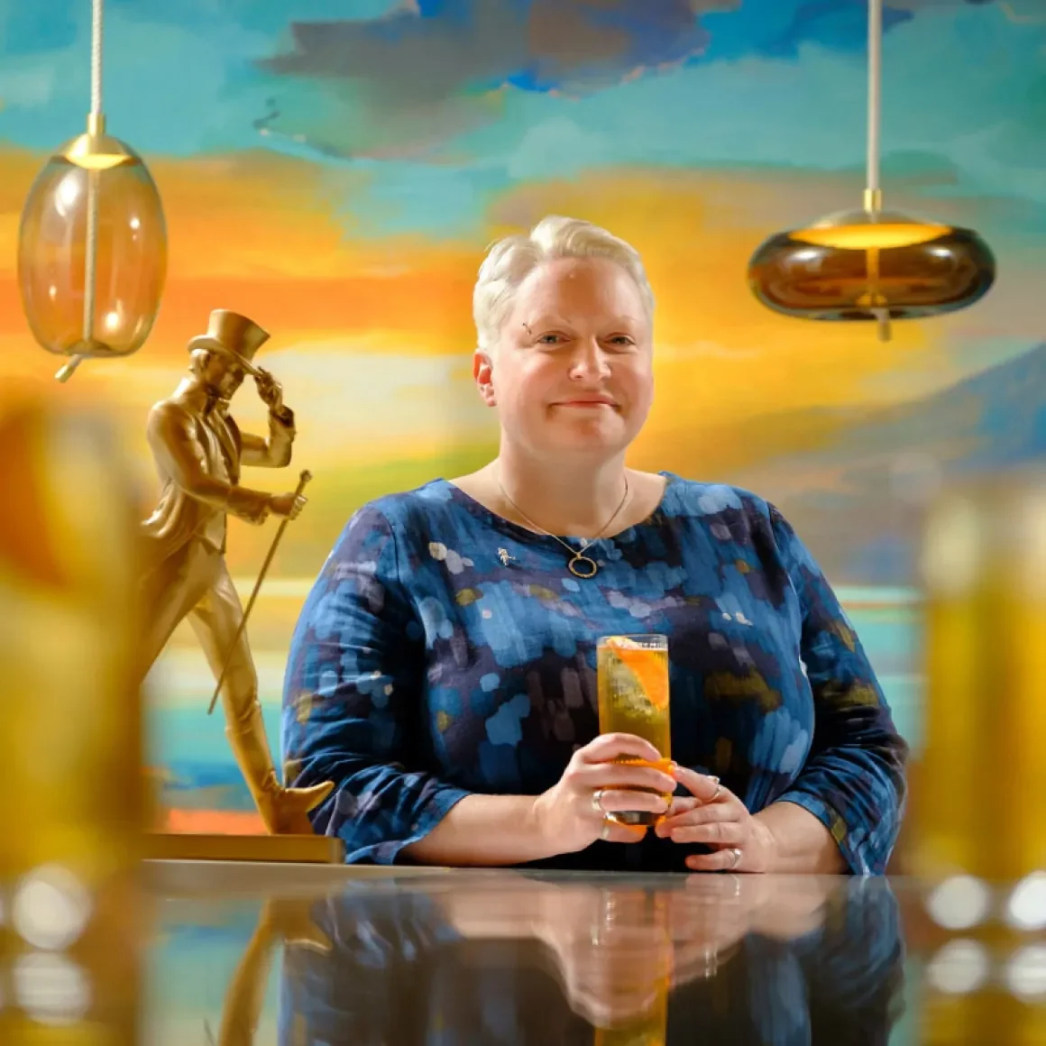 A woman with short blonde hair, wearing a blue patterned dress, stands at a bar holding a cocktail. A gold statue and pendant lights are nearby, with a colorful sunset mural in the background.