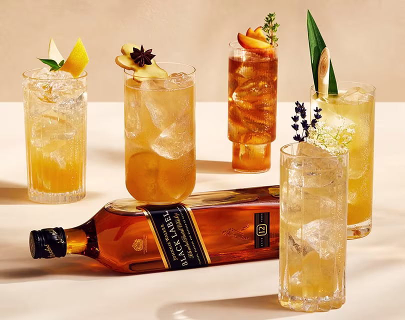 A bottle of Johnnie Walker Black Label scotch whisky lies on its side surrounded by five elegant cocktail glasses, each garnished with different fruits and herbs, on a beige background.