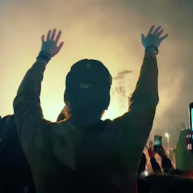 Una persona con los brazos en alto se encuentra frente a un escenario iluminado en un concierto al aire libre, rodeada de multitud. Algunos sostienen sus teléfonos para inmortalizar el momento. El ambiente es animado con una iluminación vibrante.