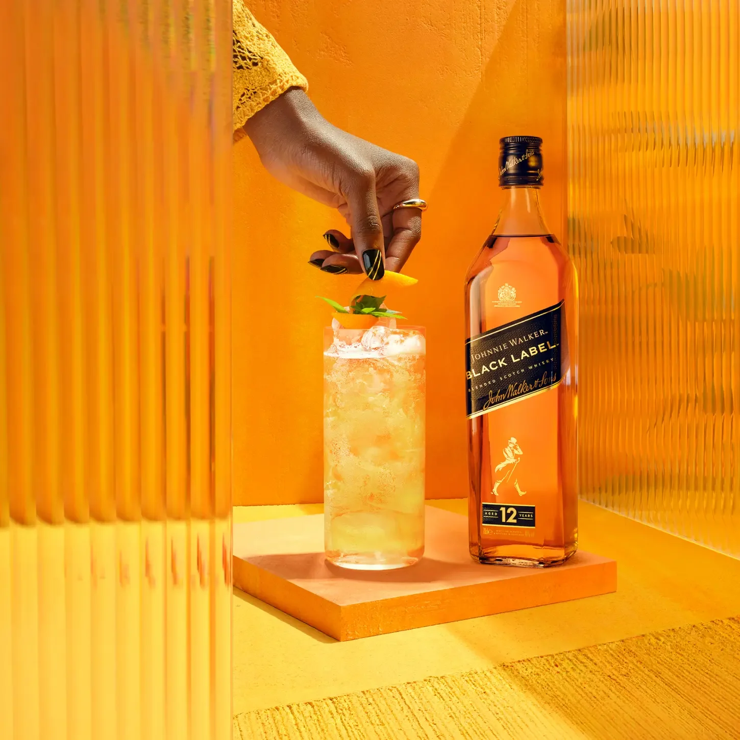 Die Hand einer Person garniert ein großes Eisgetränk mit einem Minzblatt. Das Getränk steht neben einer Flasche Johnnie Walker Black Label Whisky.