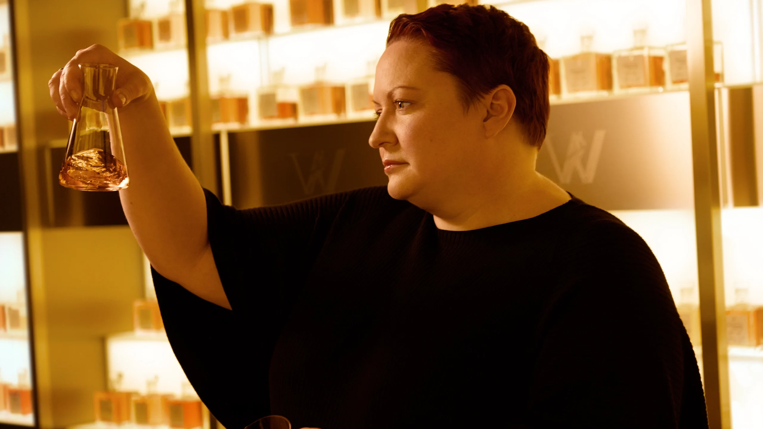 Person with short hair holds a glass container with amber liquid, examining it intently. Shelves filled with bottles are in the background, illuminated with warm lighting.