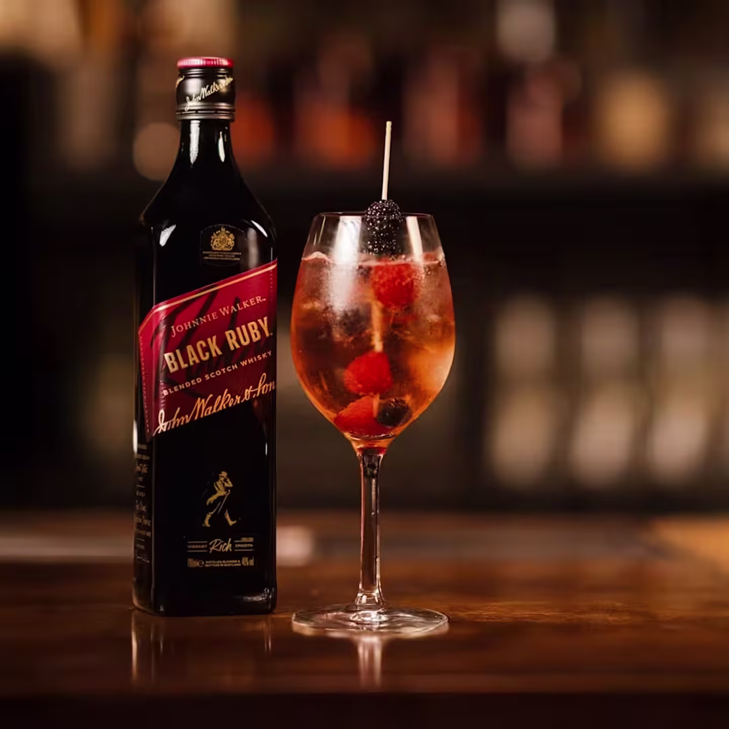 Eine Flasche Johnnie Walker Black Ruby neben einem Cocktailglas mit Eis und Beeren auf einer Bar.