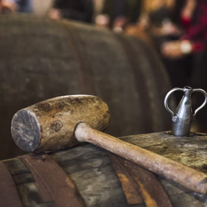 A wooden mallet and a metal spout rest on top of a wooden barrel with blurred figures in the background.