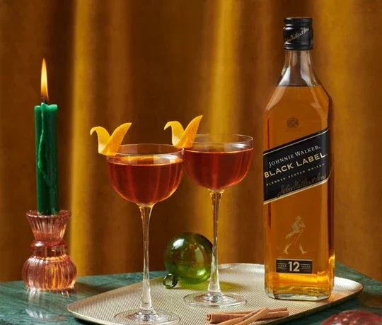 The Midnight Maple. Two cocktail glasses with orange peels sit on a table next to a bottle of Johnnie Walker Black Label. A green candle and glass ornament are in the background against a brown curtain.