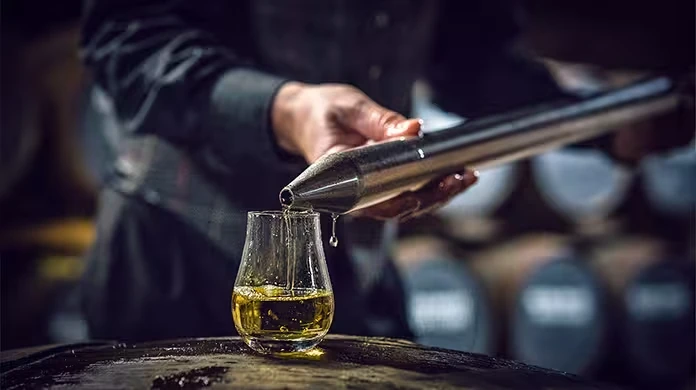 A person pours whisky from a metal tool into a glass placed on a barrel, with whisky barrels blurred in the background.