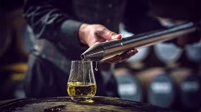 Iemand schenkt whisky met behulp van een metalen monstername-instrument in een glas. Op de achtergrond zijn de vaten vaag te zien, waardoor de nadruk ligt op een whiskyproeverij of distilleerderij.