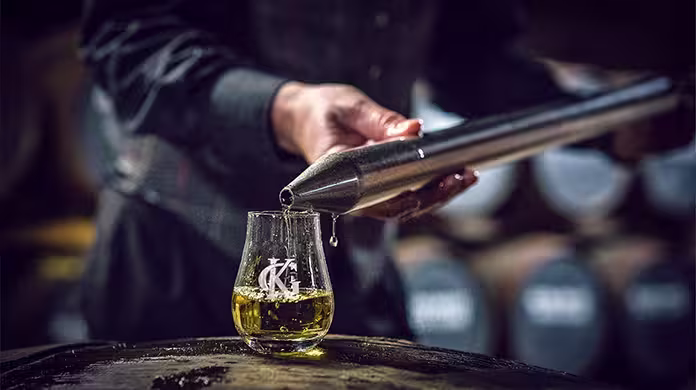 A person pours whisky from a metal tool into a glass placed on a barrel, with whisky barrels blurred in the background.