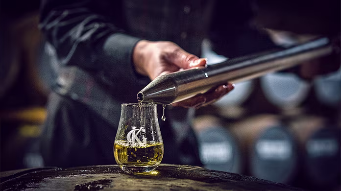 A person pours whisky from a metal tool into a glass placed on a barrel, with whisky barrels blurred in the background.