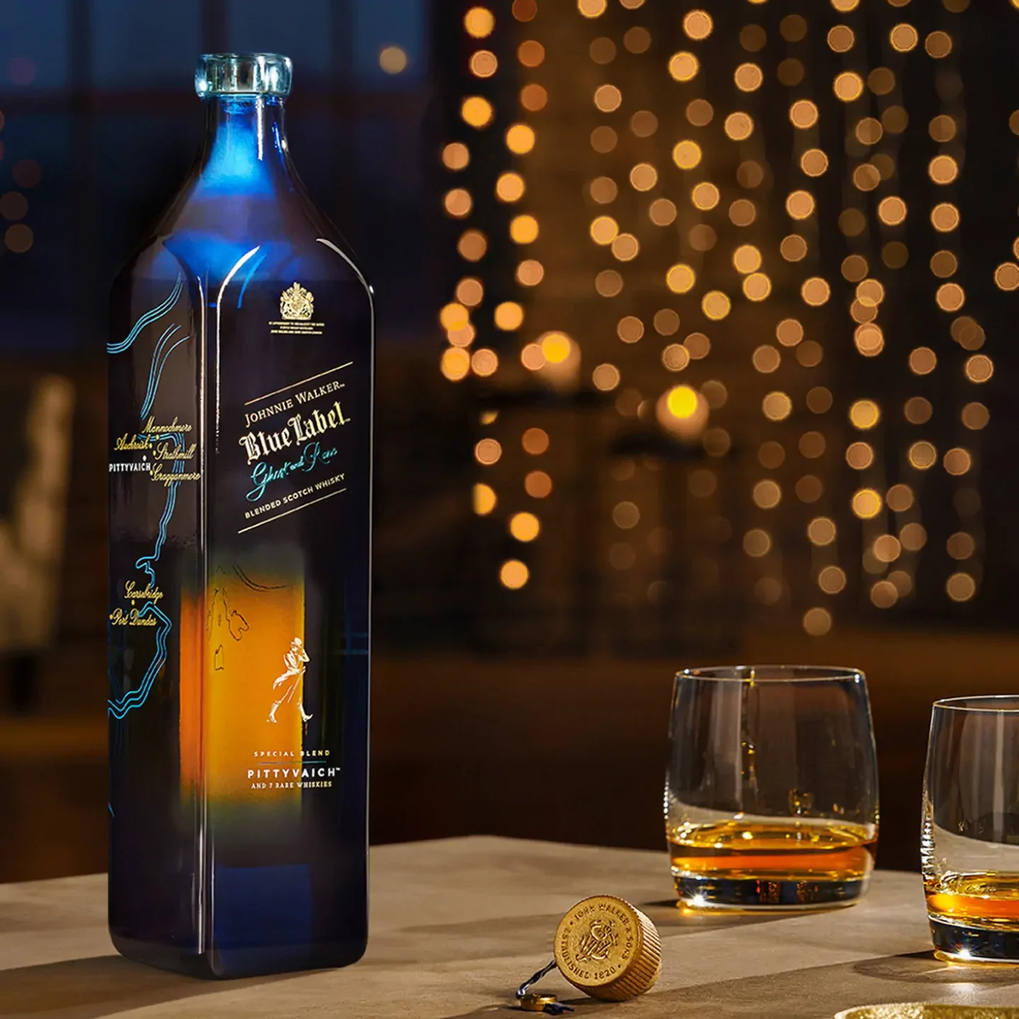 A bottle of Johnnie Walker Blue Label Ghost and Rare Pittyvaich Scotch whisky sits on a table with two glasses of whiskey and a gold bottle cap in front of a string of lights. 