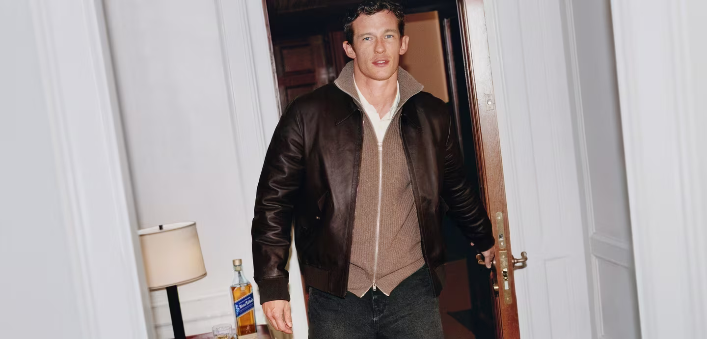 Callum Turner in a brown jacket enters a room, with a lamp and bottle on a table nearby.