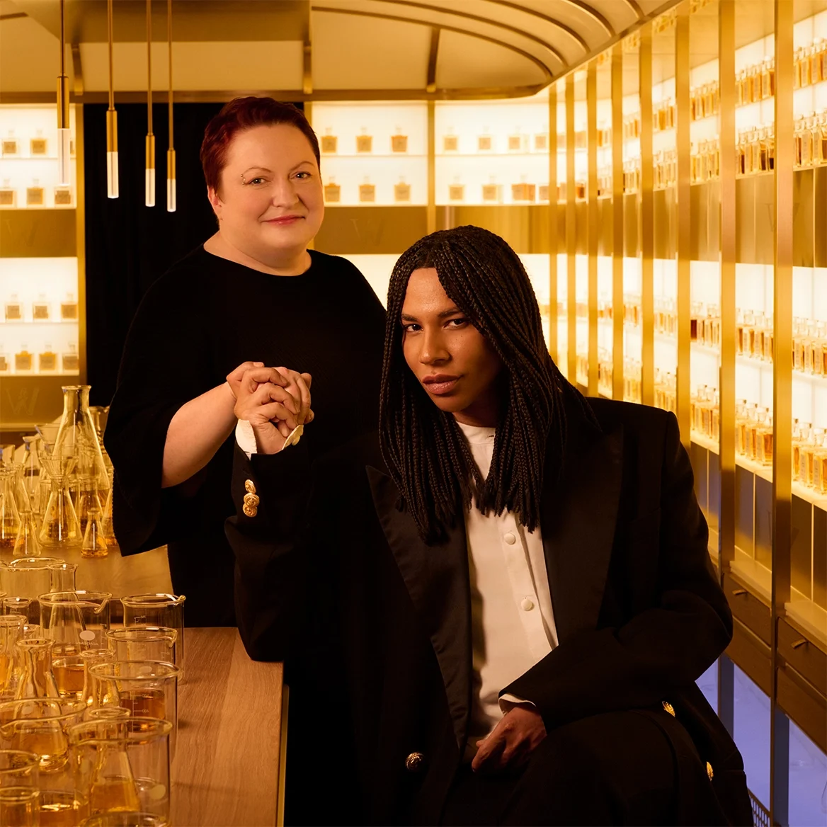 Two people pose in an elegant, warmly lit room lined with perfume bottles. One person is seated in a tuxedo with braided hair, holding hands with a standing person dressed in black and smiling, surrounded by golden glassware.