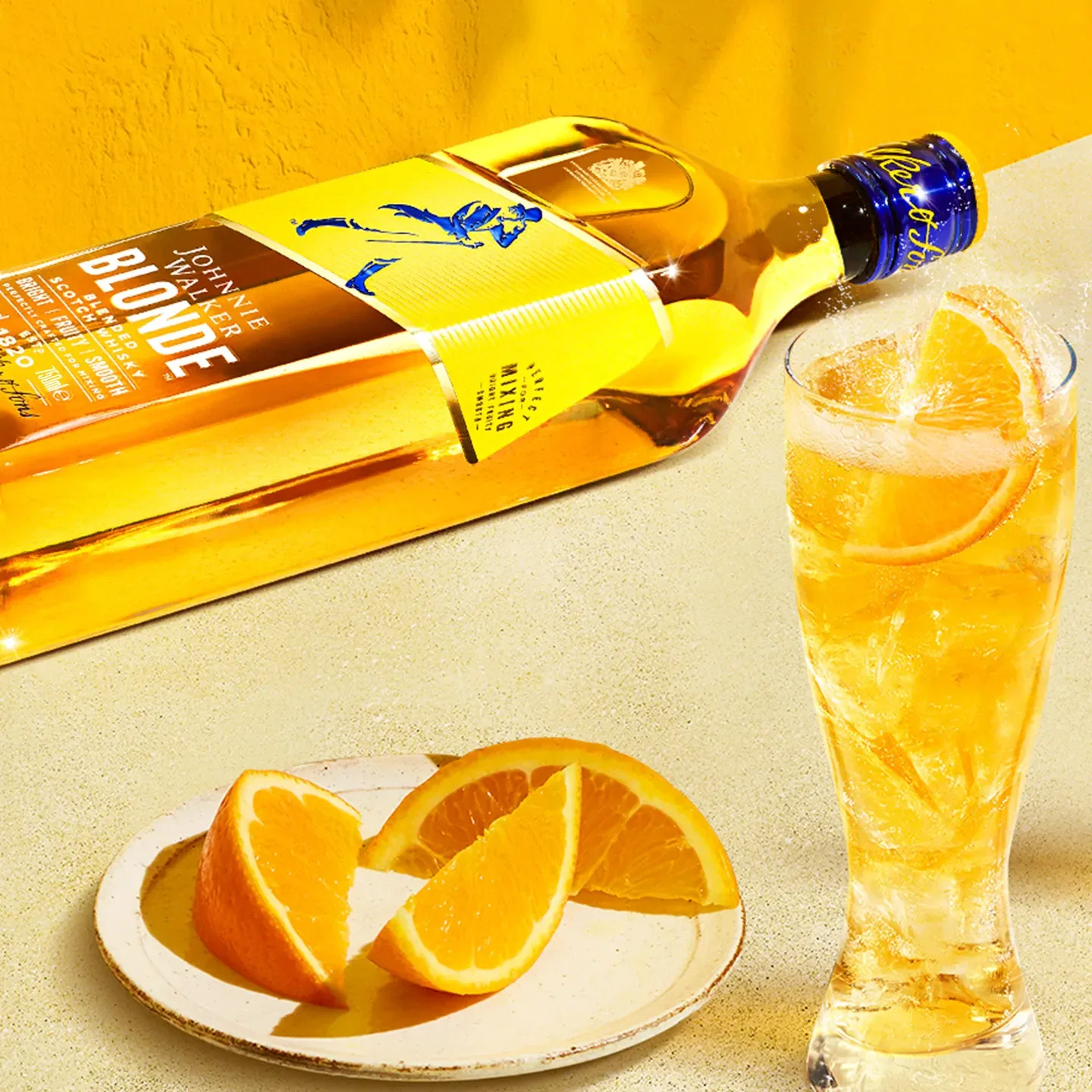 Una botella de whisky Johnnie Walker Blonde reposa sobre una superficie amarilla junto a un vaso alto lleno de una bebida de color naranja y hielo. En primer plano, sobre un fondo amarillo cálido, hay un plato con rodajas de naranja.