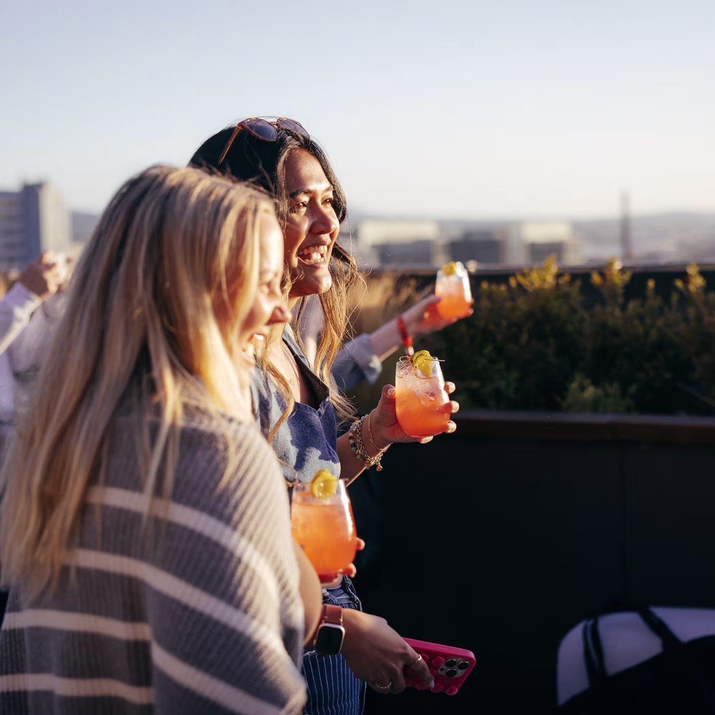 Two women holding colorful cocktails smile together on a rooftop during sunset, enjoying the view.