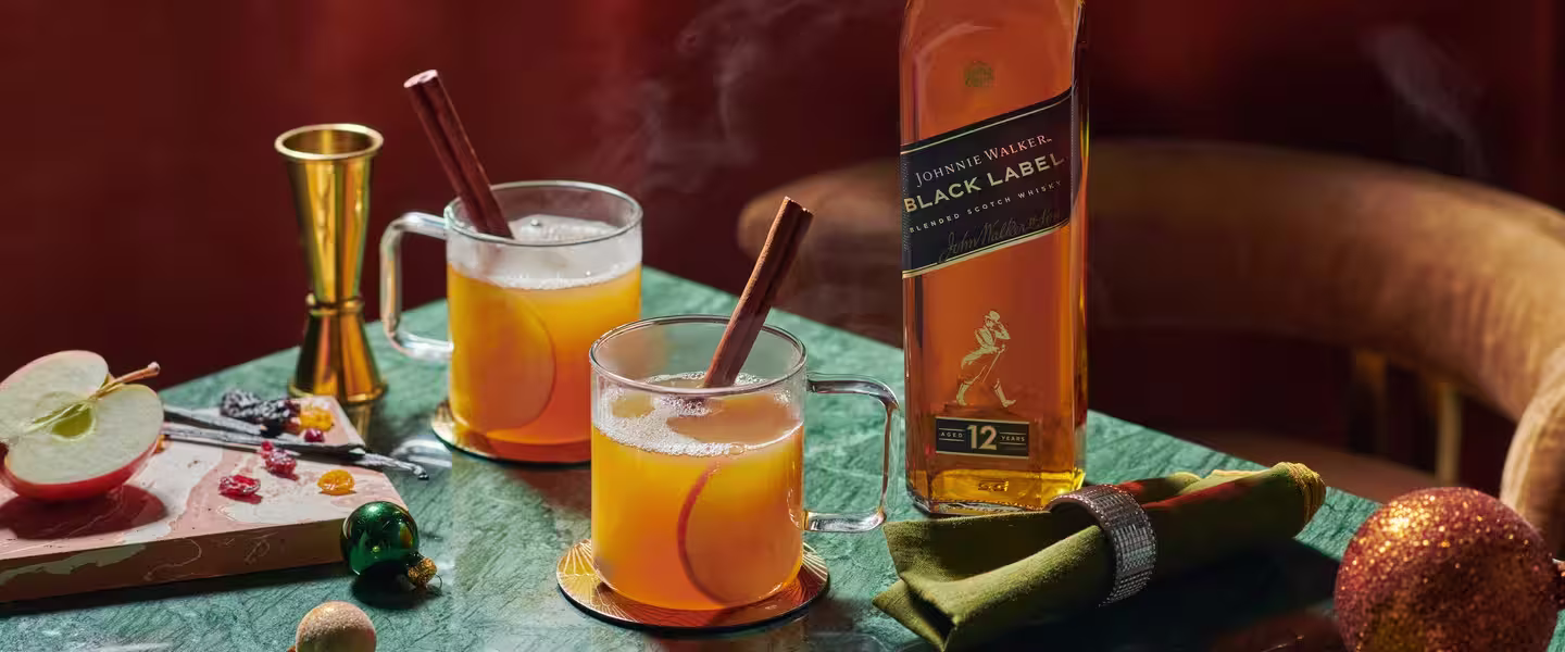 Two steaming glass mugs of a warm cocktail with cinnamon sticks sit on a green marble table. A bottle of whisky and a cut apple are nearby, along with decorative items like ornaments and a golden jigger.