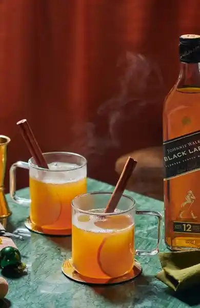 Two steaming glass mugs of a warm cocktail with cinnamon sticks sit on a green marble table. A bottle of Johnnie Walker Black Label whisky and a cut apple are nearby, along with decorative items like ornaments and a golden jigger.