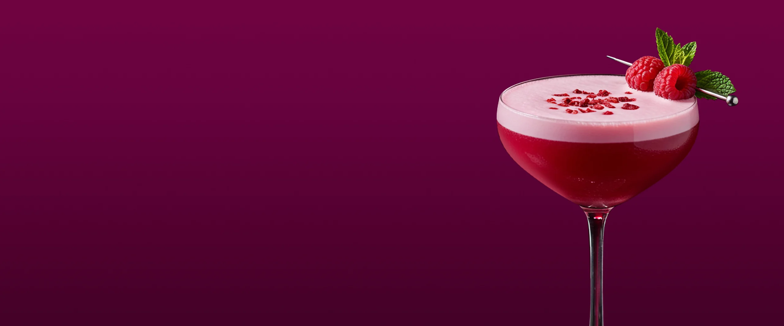 Un cóctel rosa vibrante en copa coupé sobre un fondo magenta intenso. La bebida se adorna con dos frambuesas y una ramita de menta en una brocheta, con una capa espumosa y pequeñas hojuelas rojas espolvoreadas por encima.