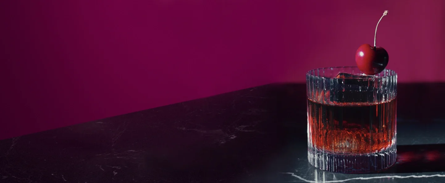 A clear glass of Black Ruby Negroni with a large ice cube sits on a dark, glossy surface. A cherry garnishes the drink. The background is a deep magenta colour, creating a rich and elegant atmosphere.