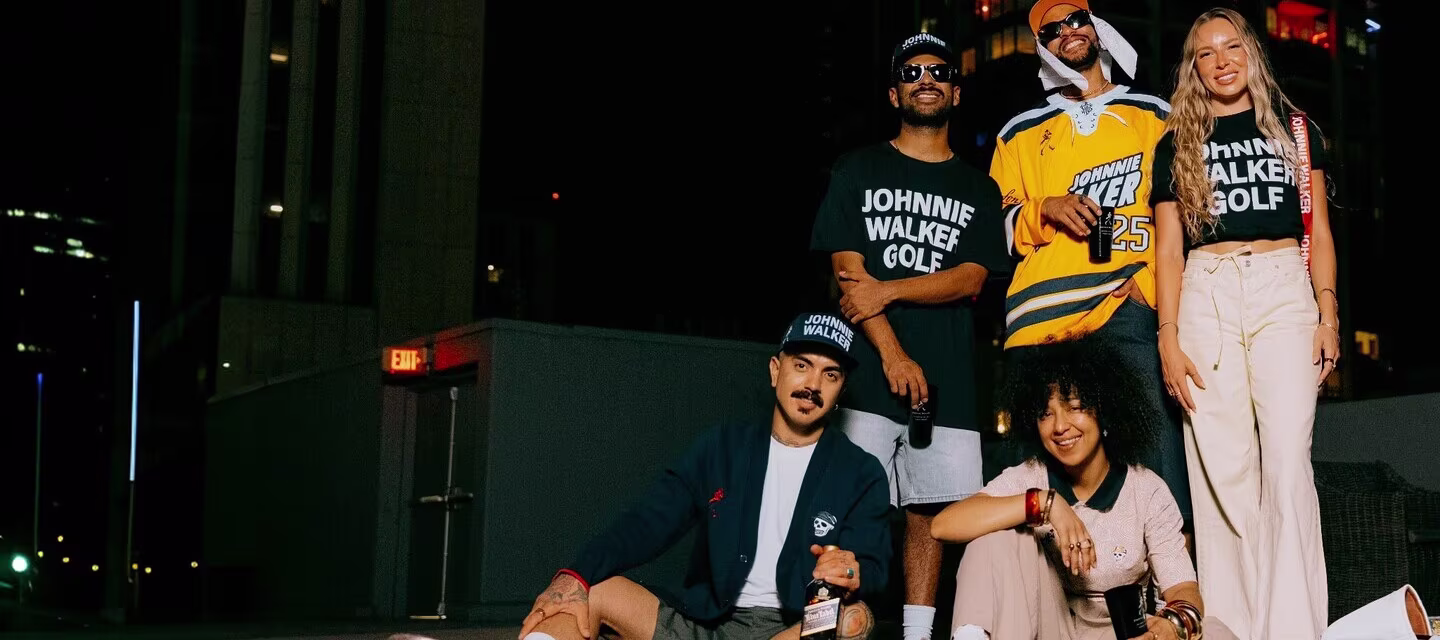 Five young adults pose together on a rooftop at night, some standing and others sitting on the grass. They wear casual clothing, including shirts with JOHNNIE WALKER GOLF, and two have bottles in hand. City buildings are visible in the background.