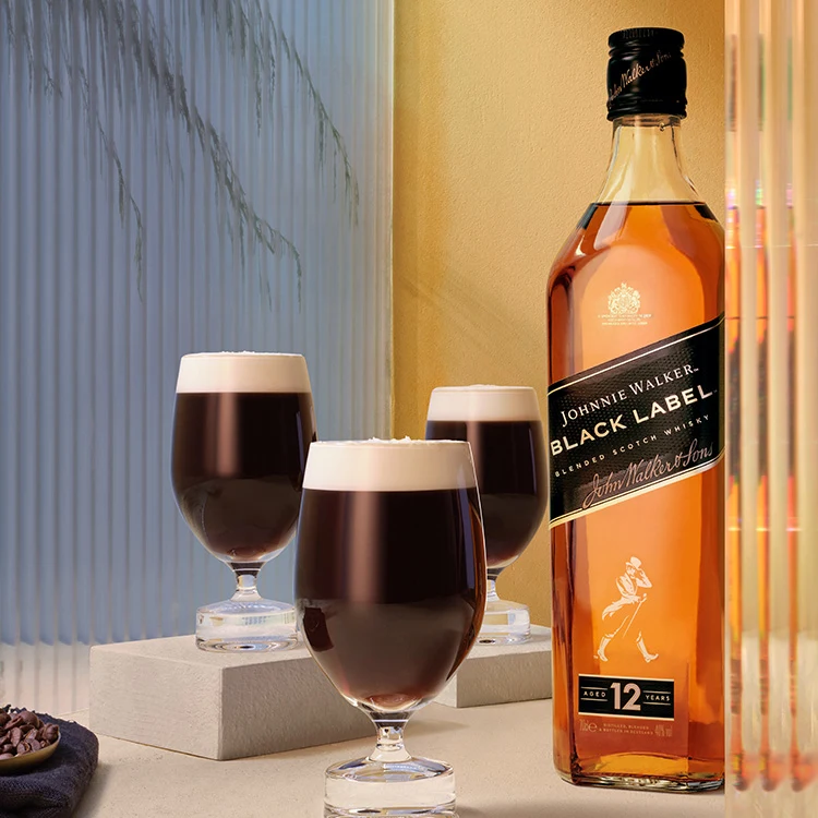 A bottle of Johnnie Walker Black Label sits next to three glasses of a dark cocktail with creamy tops. The bottle and drinks are placed on a beige surface near a lightly textured wall, with soft lighting creating a cosy atmosphere.